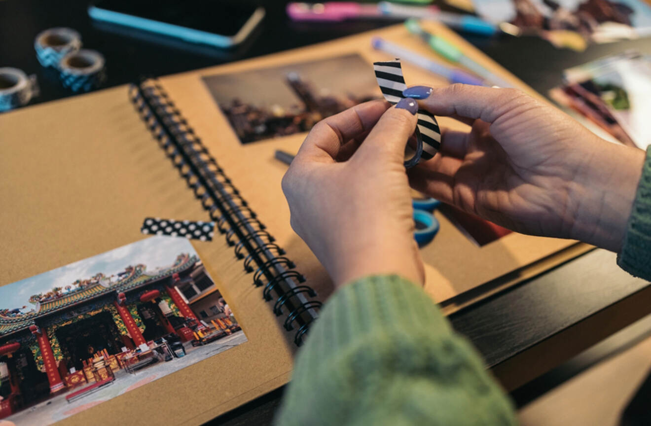 Hands decorating a travel scrapbook with photos and decorative tape.