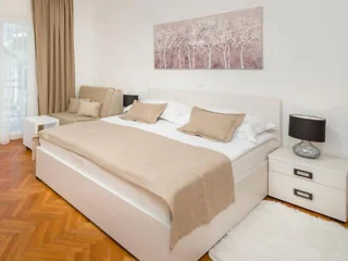 A bedroom with a large bed covered in beige linens, flanked by matching nightstands with black lamps. A beige couch is in the corner, and a tree-themed artwork hangs above the bed.