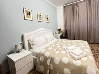 A bedroom with a double bed, polka dot beige bedspread, bedside table with a lamp, and a desk with a chair in the corner. Framed pictures hang above the bed and large curtains cover the window.