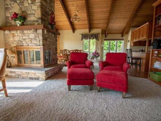 A cozy living room features a stone fireplace, two red armchairs with matching ottomans, a dining area in the background, and a high wooden ceiling with exposed beams.