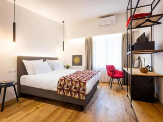 A modern bedroom with a king-size bed, a red chair, a wooden floor, a wall-mounted TV, an open wardrobe, and a work desk with a lamp.