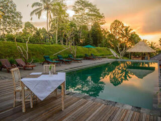 A serene outdoor pool area with lounge chairs, umbrellas, and a thatched-roof hut, set against a backdrop of lush greenery and a colorful sunset. A table with a white cloth is in the foreground.