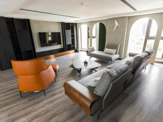 A modern living room with a large TV, gray sofas, an orange armchair, a unique coffee table, and floor-to-ceiling windows letting in natural light.