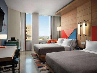 A hotel room with two double beds, a desk, chairs, a wall-mounted TV, and a window overlooking city buildings. The decor includes colorful geometric patterns and modern lighting.