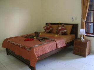 A bedroom with a double bed dressed in brown and red linens, two pillows, a towel sculpture on the bed, a bedside table with a lamp, and a window with drawn curtains.