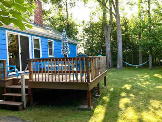 A blue house with a wooden deck, outdoor furniture, and a striped umbrella set in a grassy backyard with trees and a hammock in the background.