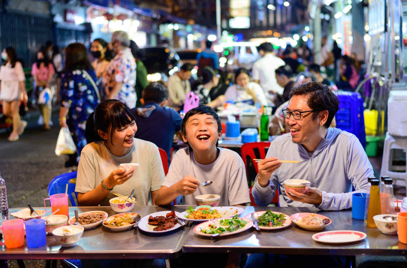 Family laughing and sharing a meal at a bustling night market in Bangkok, Thailand.
