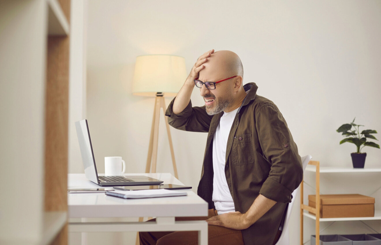 A man in a home office laughs while looking at his laptop, holding his head in amusement.