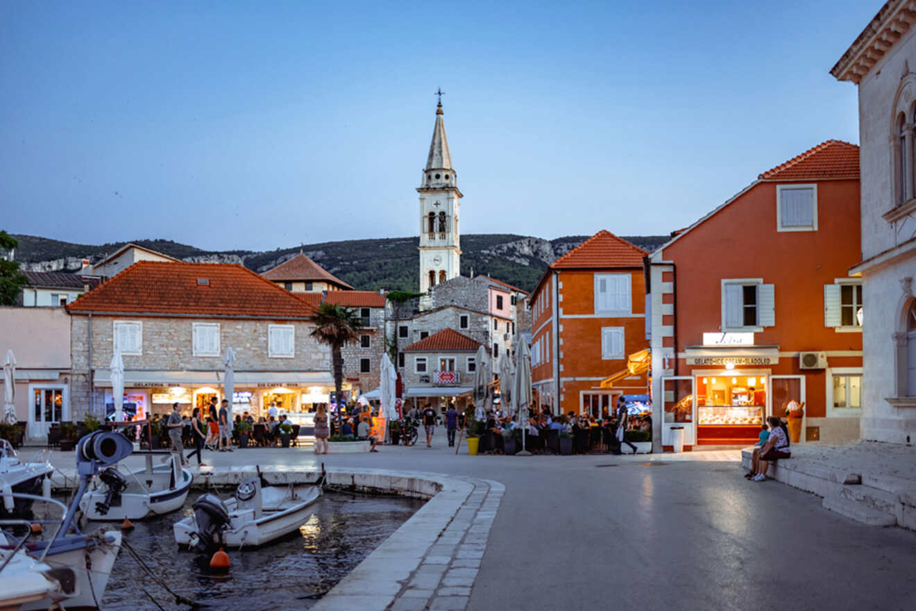 A picturesque coastal town with brightly colored buildings, a church steeple, outdoor cafes, and docked boats at dusk.