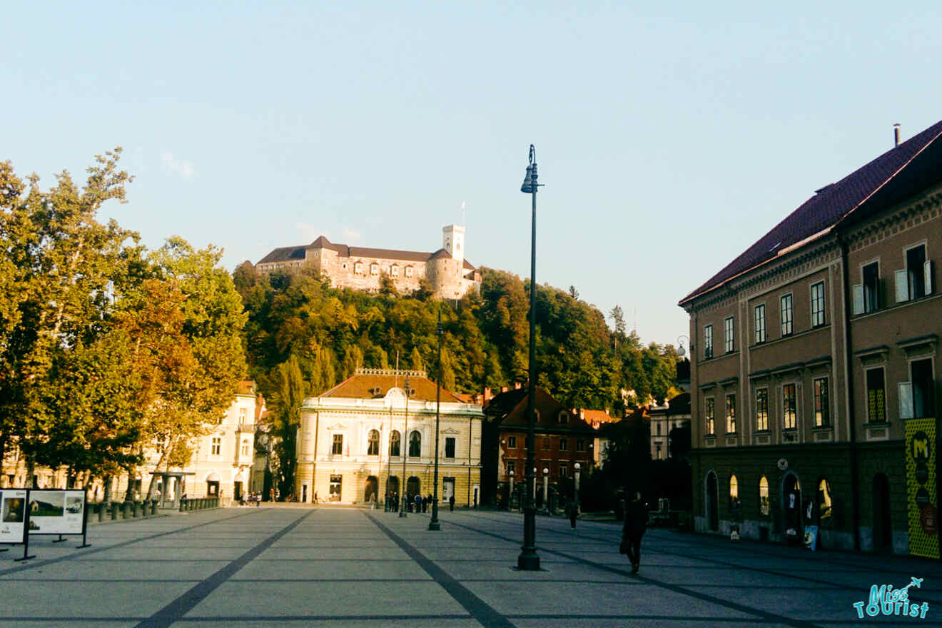 A spacious town square with trees and historic buildings, under a clear sky. A castle stands on a hill in the background.