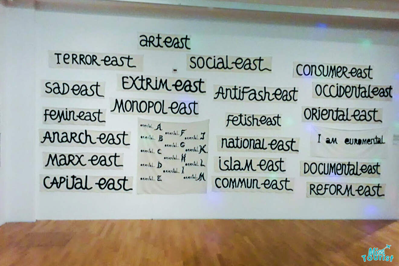 A white wall covered with various handwritten terms ending in "-east," such as "arteast," "sad east," "antifash east," and "consumer east," among others, on white paper sheets.