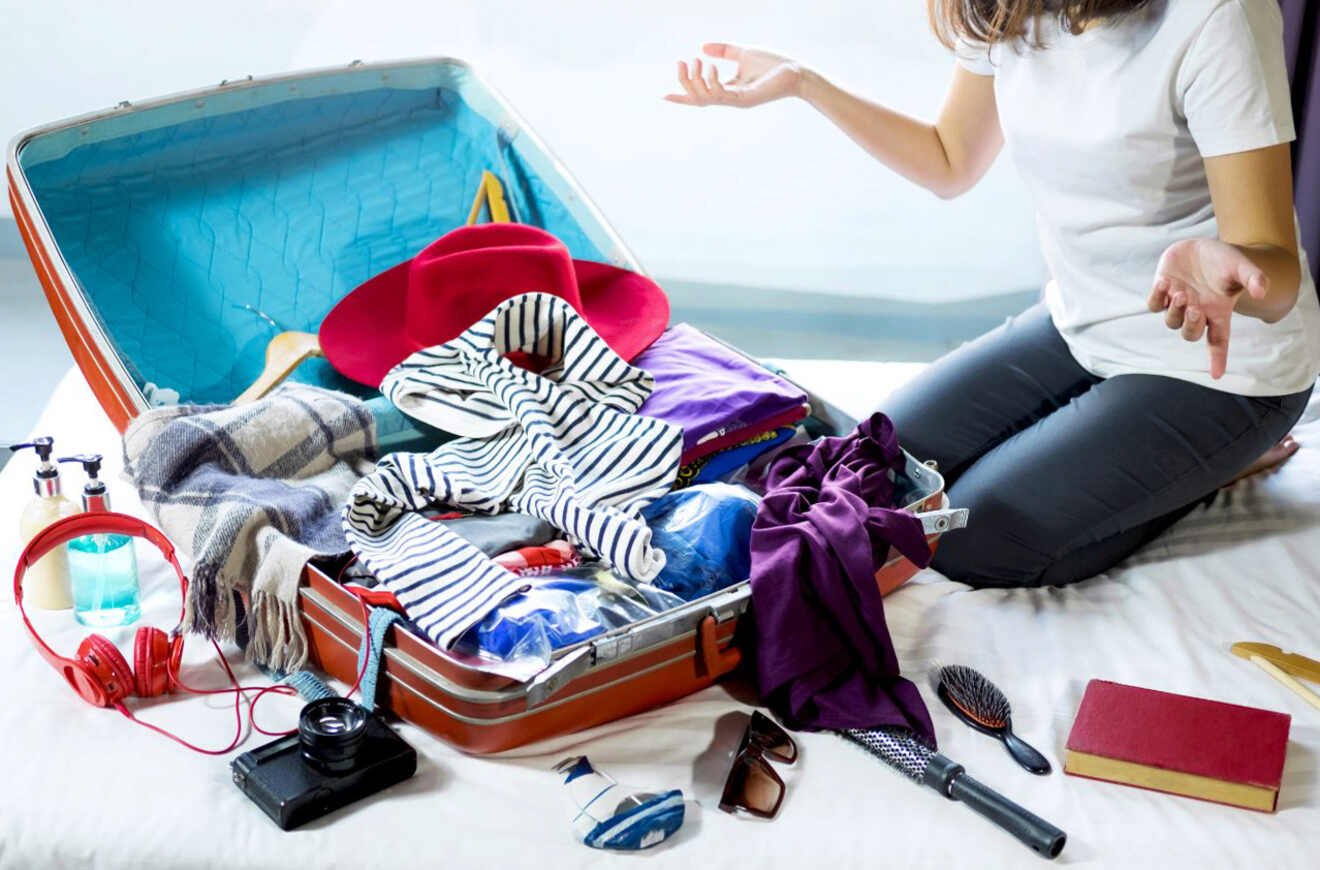 An open suitcase overflowing with clothes, including a red hat, striped shirt, and various travel items, with a person sitting next to it looking frustrated
