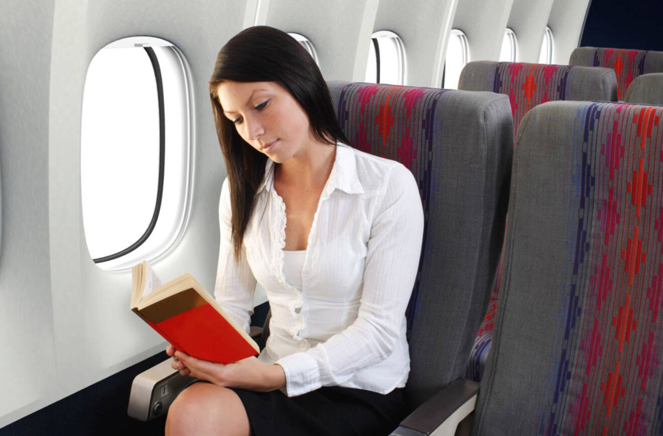 A woman with long dark hair reads a book while seated on an airplane with patterned seats.