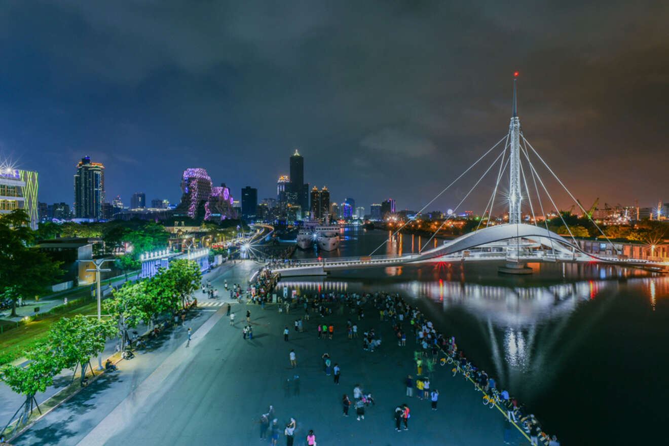 Night view of a cityscape featuring a river, a modern pedestrian bridge with cable supports, and numerous people gathered on a plaza. The skyline is illuminated with colorful lights.