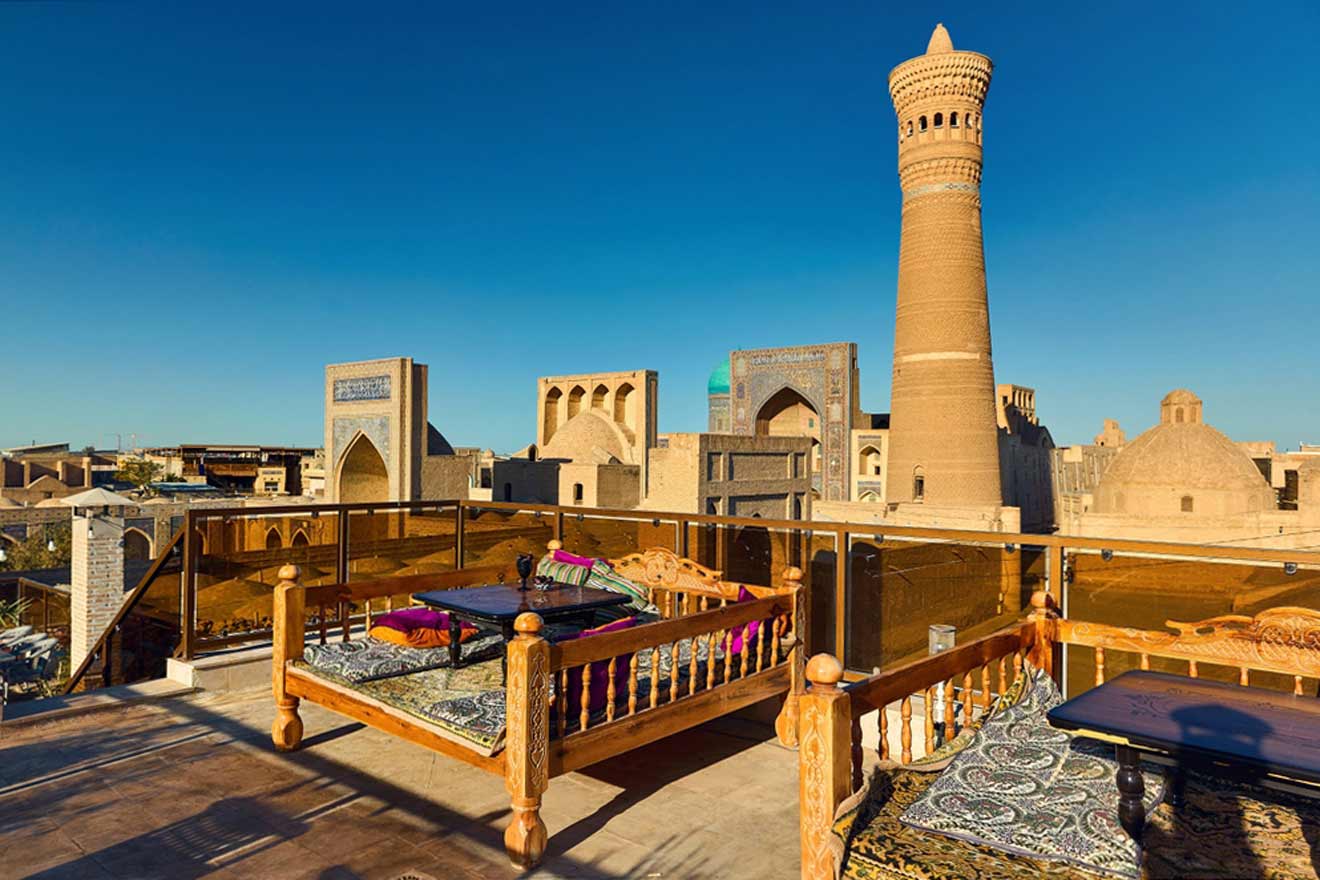 A rooftop terrace with traditional wooden furniture overlooks ancient architecture and a tall minaret in a historic city under a clear blue sky.