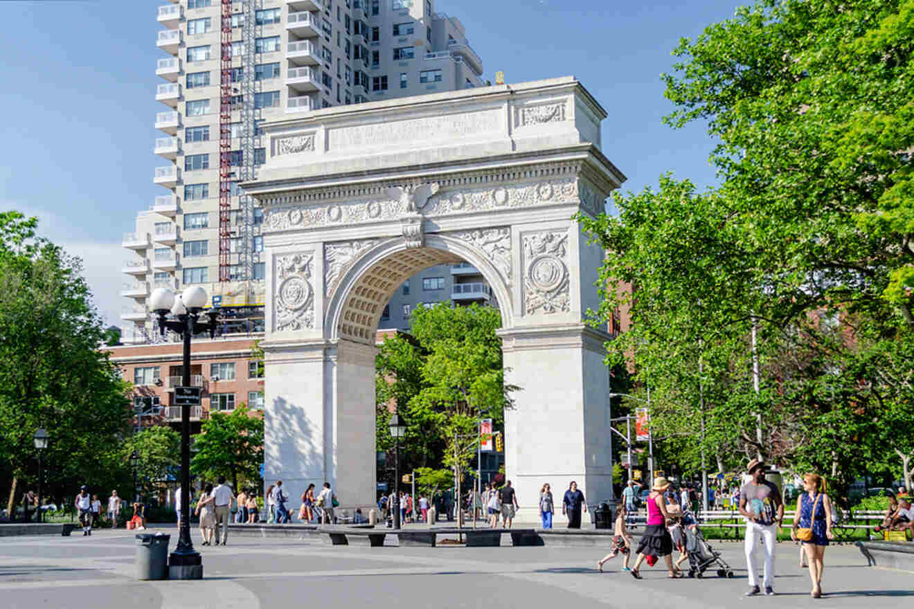 People stroll through a park with an ornate arch and tall buildings in the background on a sunny day.