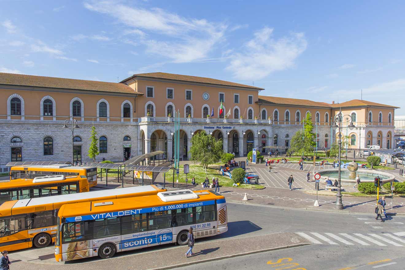Image of Pisa Centrale railway station in Italy with a plaza in front, where orange buses are parked, people walking, and a fountain visible. The station has an arched facade and several flags flying.