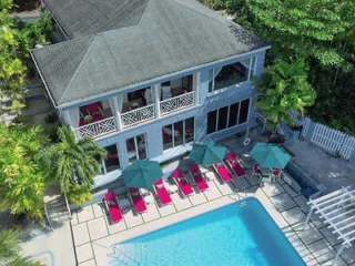 Aerial view of a two-story house with a large outdoor swimming pool, several red lounge chairs, and green umbrellas on a patio surrounded by trees.