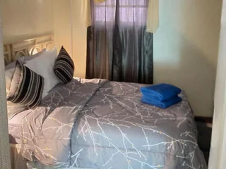 A small bedroom with a double bed featuring a gray and white patterned comforter, striped pillows, and a stack of blue towels on the bed. The room has a window with sheer and dark curtains.