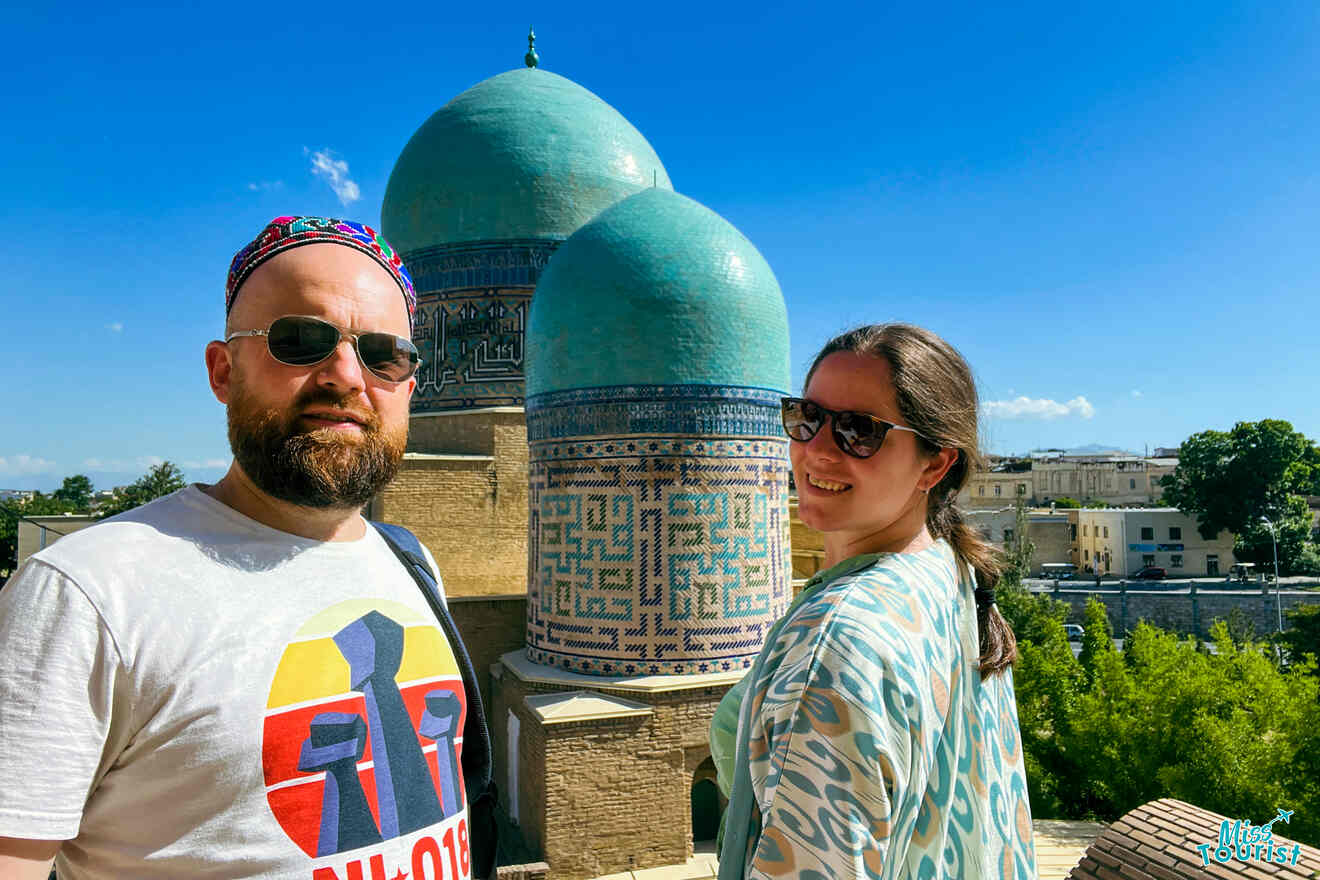 author of the post with husband wearing sunglasses stand in front of blue-domed buildings with intricate mosaic designs under a clear sky. One person wears a graphic T-shirt and a patterned hat, while the other wears a patterned jacket.