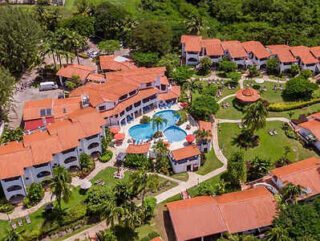 An aerial view of a resort featuring red-roofed buildings, a central swimming pool, pathways, green lawns, and palm trees.