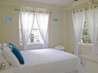 A bright bedroom with white walls and curtains, a double bed with white linens and blue accent pillows, and two windows allowing natural light to enter. An air conditioner is installed above one window.