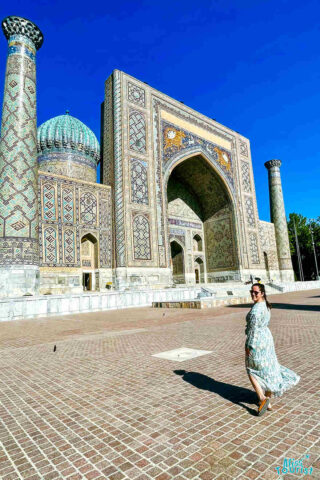 author of the post wearing a patterned dress stands on a plaza in front of an ornate building with intricate tilework and blue domes under a clear sky.