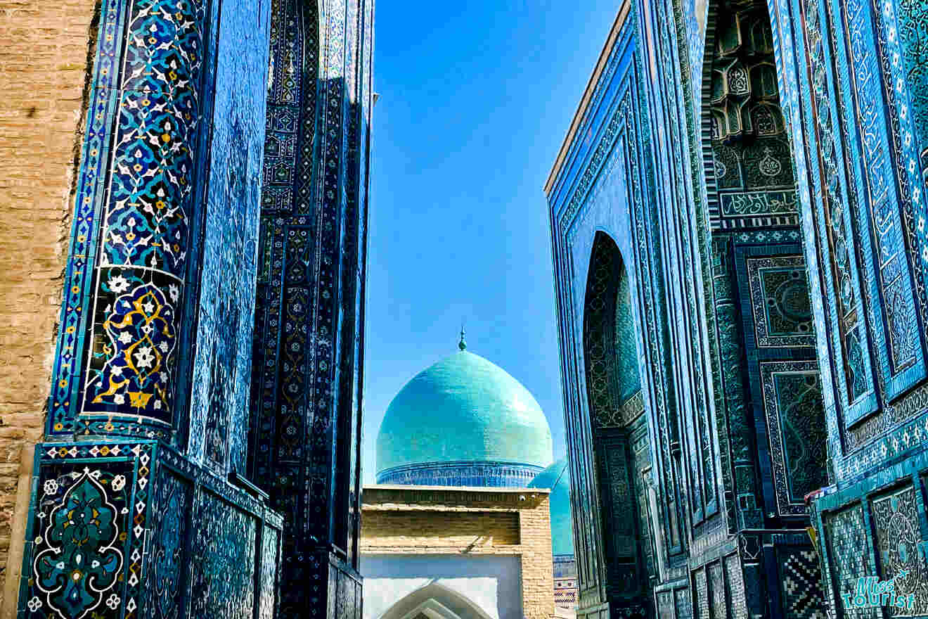 A pathway lined with intricate tiled walls leads to a structure with a large turquoise dome in an architectural complex under a clear blue sky.