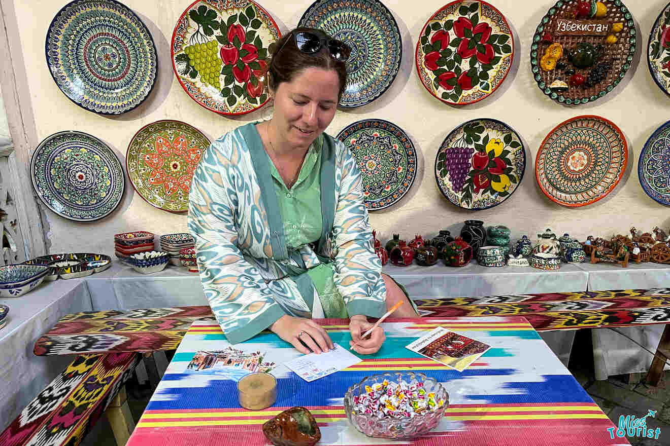 author of the post in a green robe stands at a colorful table, working with small papers. Decorative plates with intricate patterns are displayed on the wall behind her in a shop.