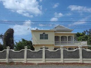 A two-story beige house with a decorative balcony, surrounded by a high stone and concrete fence, set against a partly cloudy sky.