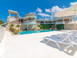 A large, white, multi-story villa with arched balconies and a private pool surrounded by lounge chairs, under a clear blue sky.