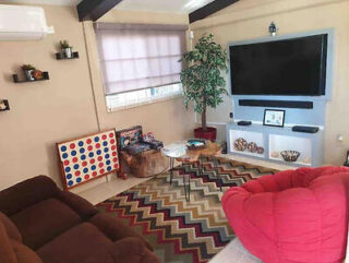 A living room with a large TV on a wall-mounted unit, a sofa, a red bean bag chair, a patterned rug, potted plants, and a Connect Four game set up near the window.