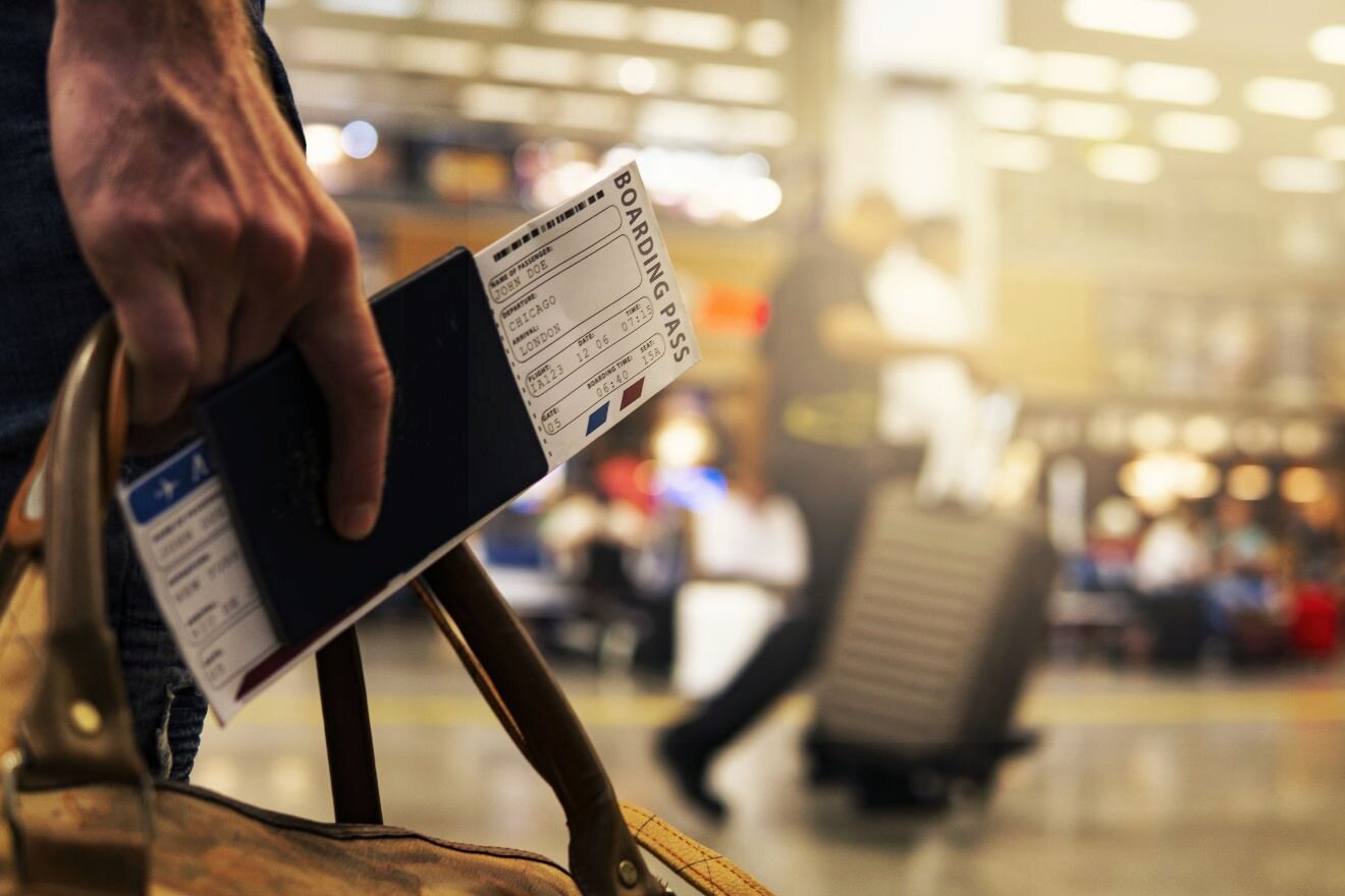 A hand holding a passport with a boarding pass, standing in an airport terminal with a blurred background of travelers and luggage.