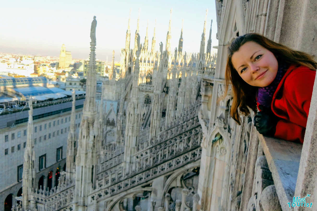 Yulia, la fundadora de este sitio web, con un abrigo rojo mira desde un balcón de una gran catedral gótica ornamentada con intrincadas agujas y un paisaje urbano visible en el fondo.