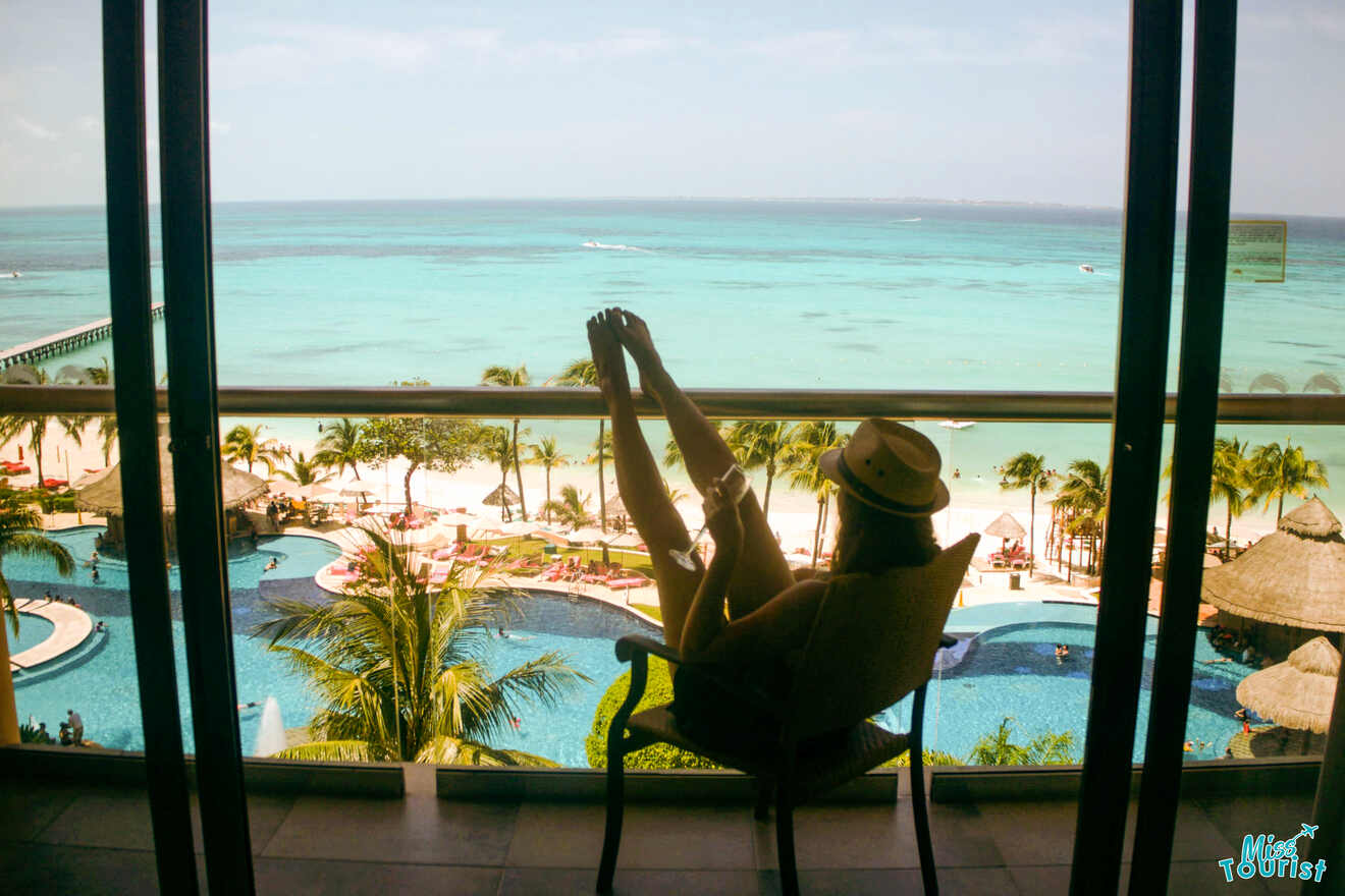Yulia, la fundadora de este sitio web, se relaja en un balcón con vista a una playa tropical con aguas turquesas cristalinas, palmeras y una piscina.