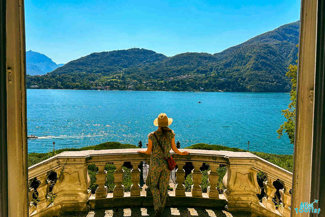 Yulia, la fundadora de este sitio web, con sombrero, se encuentra en un balcón de piedra con vistas a un gran lago rodeado de colinas bajo un cielo azul claro.