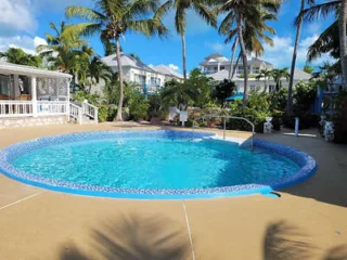 A round outdoor pool with clear blue water surrounded by palm trees, a white building, and sunny skies in a tropical setting.