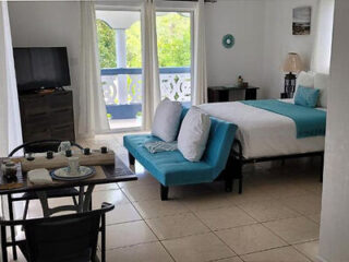 A compact vacation rental room with a bed, turquoise sofa, dining table, TV, and a balcony with green views. The room has white walls with minimal decorations and tiled flooring.