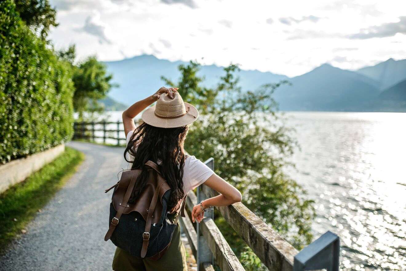 A person with a hat and backpack stands by a wooden fence along a lake, with mountains in the distance on a cloudy day.