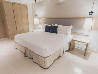 A neatly made bed with white linens and two navy blue pillows in a modern bedroom with beige walls and a wooden closet. Two bedside tables with lamps are on either side of the bed.