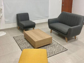 A living room with gray sofa and armchair, yellow ottoman, wooden coffee table on patterned rug, and a window with a closed blind.