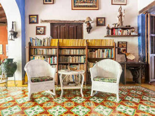 Two white wicker chairs with cushions and a small table sit in front of a bookshelf filled with books in a colorful room with patterned floor tiles and various wall decorations.