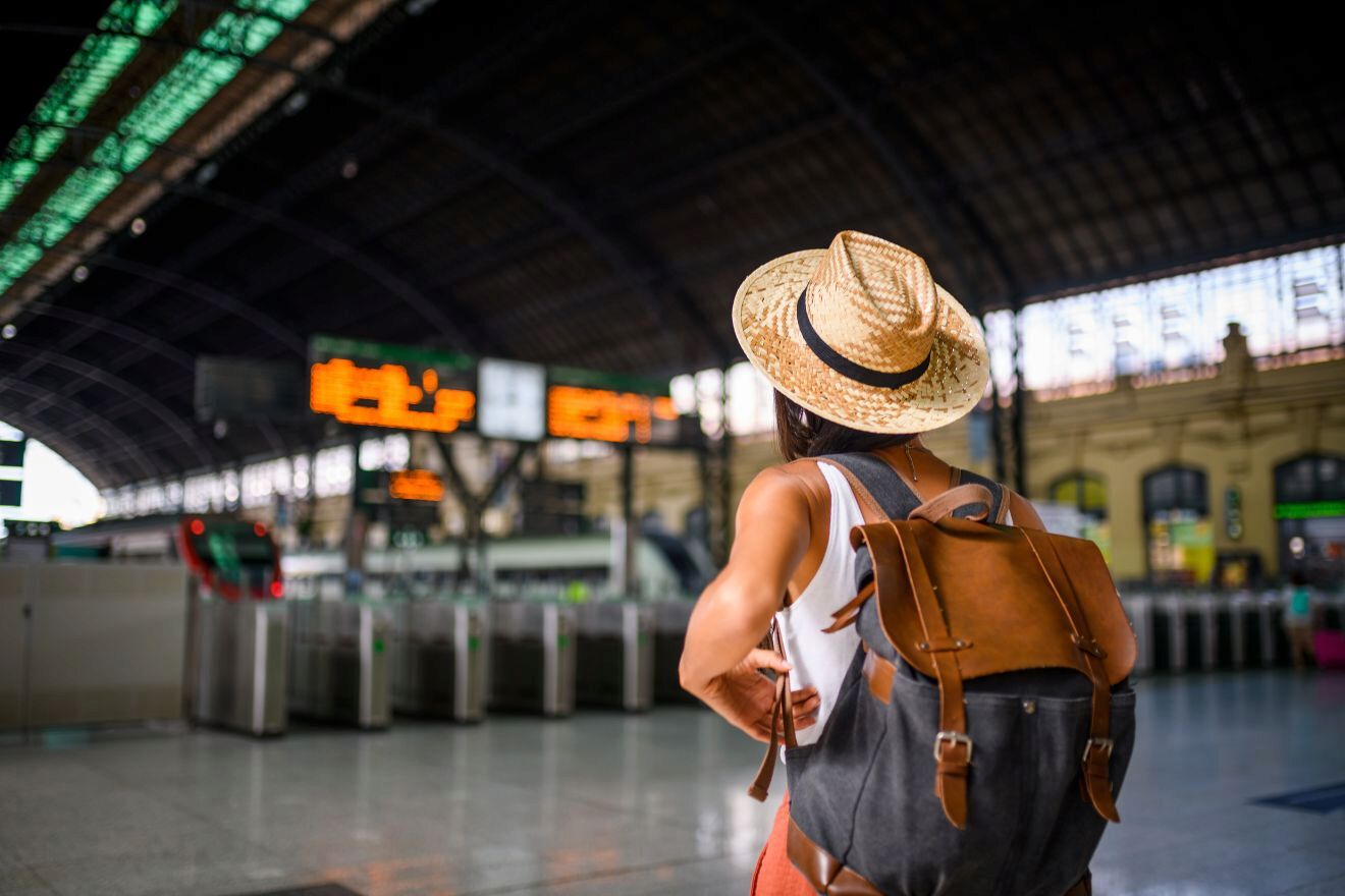 A traveler wearing a straw hat and backpack stands in a train station, looking at the electronic schedule board.