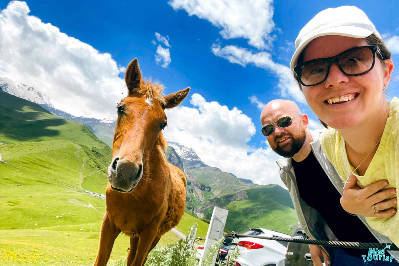 Darija Stojanovic, la autora de las publicaciones de misstourist, con su esposo, posa para un selfie con un caballo marrón en un paisaje montañoso verde en un día soleado.