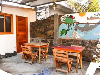 Outdoor seating area with two wooden tables and four chairs under a corrugated metal roof. A mural with sea creatures decorates the stone wall. A wooden door and window are visible.