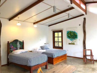 A bedroom with two single beds, patterned bedding, and a wooden headboard. The room features white walls, wooden beams, a window, hanging light fixtures, a wooden chair, and nature-themed artwork.