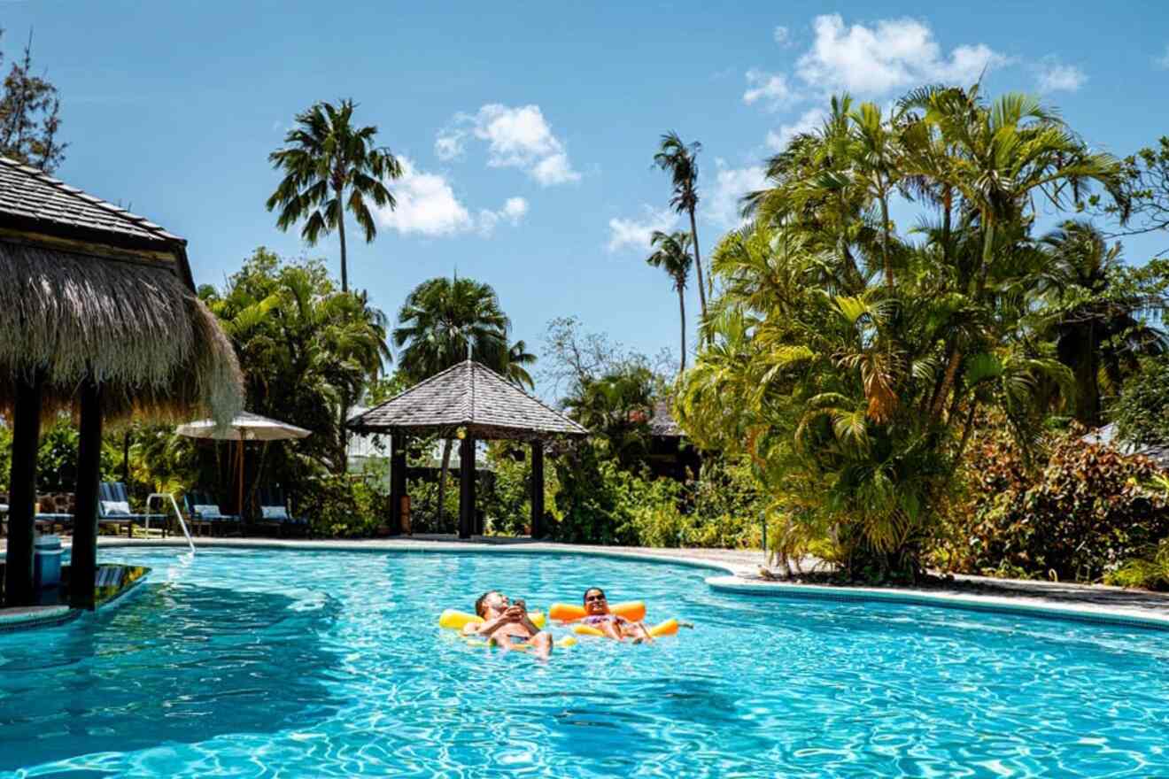 Two people relax on inflatable floats in a resort swimming pool surrounded by lush tropical vegetation and thatched structures under a clear blue sky.