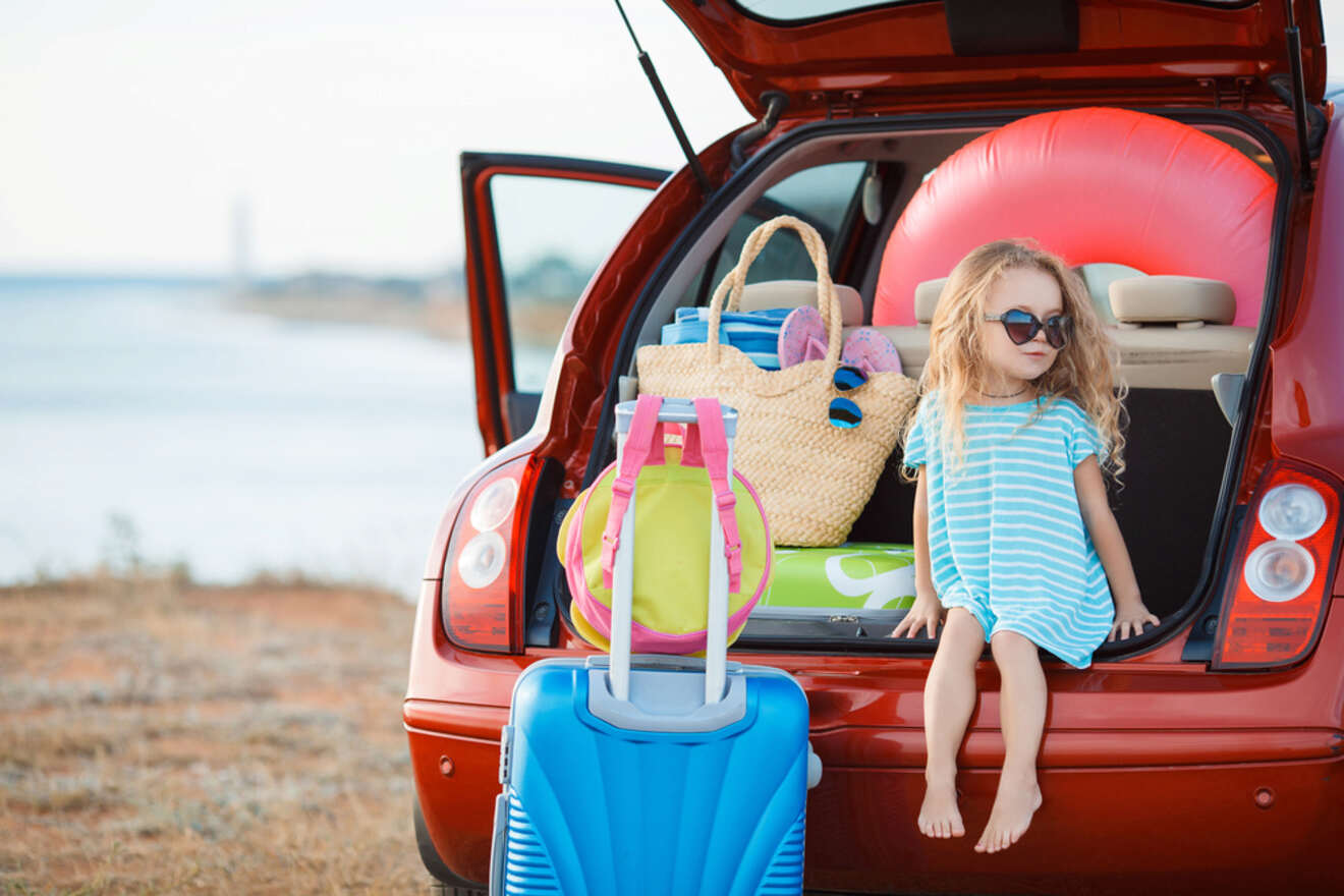 A young girl wearing sunglasses sits at the back of a red car packed with beach items, including a large inflatable ring, a straw bag, and a suitcase, with a beach scene in the background.
