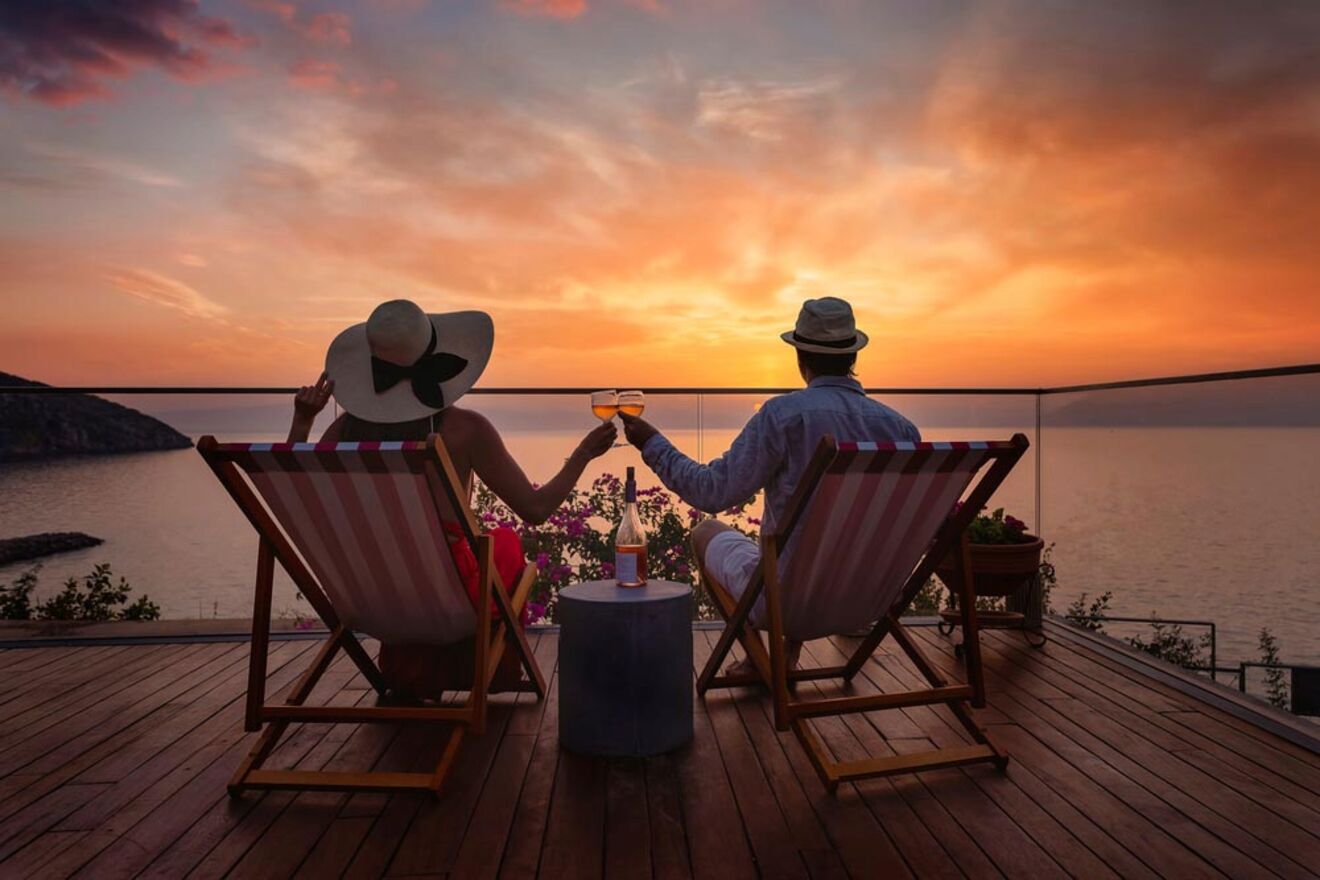 Two people sit in deck chairs on a wooden terrace, holding hands while facing a sunset over the ocean. They each hold a glass of wine with a bottle on a small table between them.