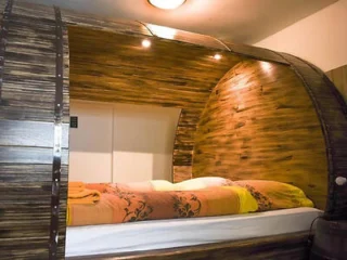 A cozy bed inside a wooden barrel-shaped structure with a patterned orange and yellow bedspread, illuminated by a ceiling light.